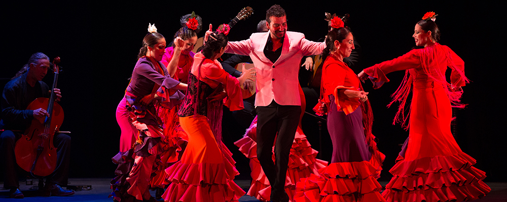 dancers in long red spanish-style dresses surround a man dancing in a white suit jacket with black pants
